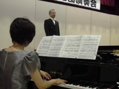 演奏会のリハーサル風景。男性(1名)が壇上で歌い、女性(1名)ピアノを演奏している様子