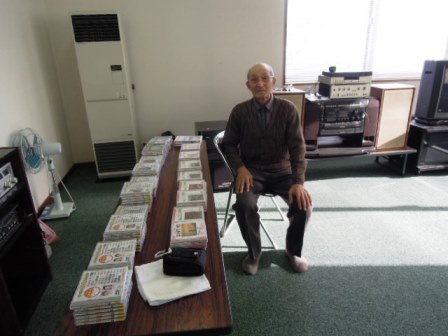 カラオケ担当の竹本さんがイスに座っており、前の机にはたくさんのCDが並んでいます。