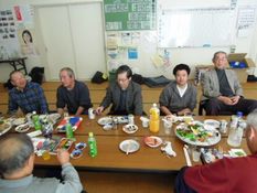 新年会の様子を写した画像。男性が7名座っており、食事や談笑をしています。
